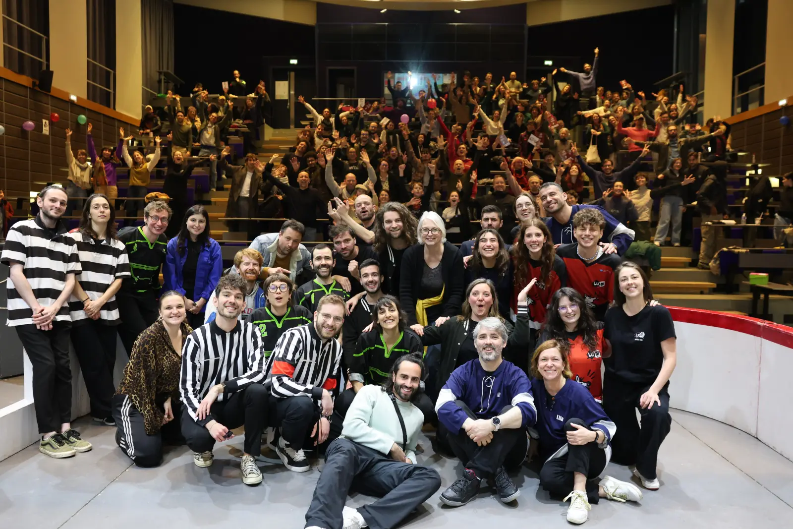 Photo de groupe des joueuses et joueurs de la LUDI sur scène, devant un amphithéâtre plein qui lève les bras